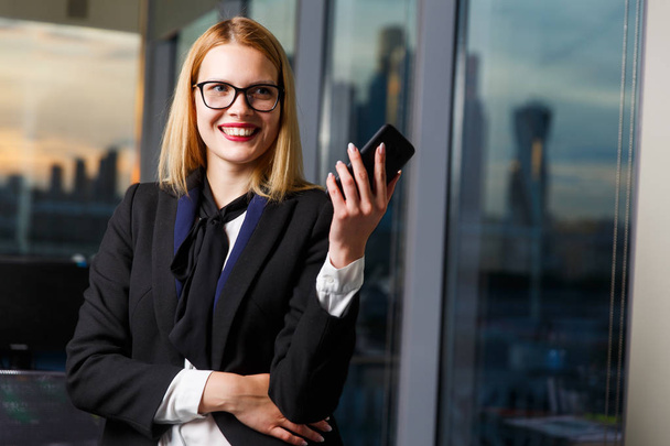 Photo of smiling woman in glasses with phone in hands near glass wall - Foto, Bild