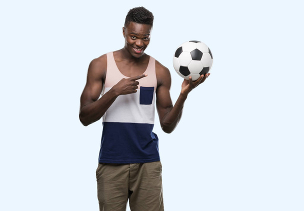 Jonge african american man die voetbal voetbal erg blij met de hand en de vinger te wijzen - Foto, afbeelding