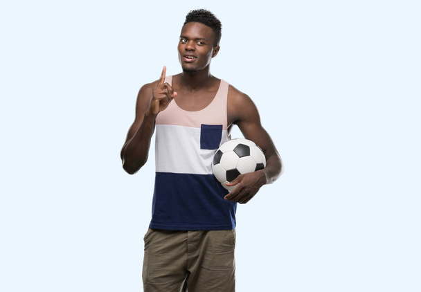 Jeune homme afro-américain tenant ballon de football surpris par une idée ou une question pointant du doigt avec un visage heureux, numéro un
 - Photo, image