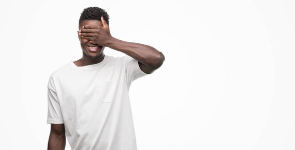 Joven hombre afroamericano vistiendo camiseta blanca sonriendo y riendo con la mano en la cara cubriendo los ojos para sorpresa. Concepto ciego
. - Foto, Imagen