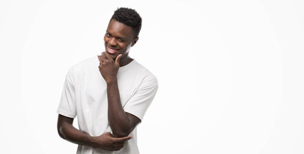 Jeune homme afro-américain portant un t-shirt blanc regardant avec confiance la caméra avec le sourire avec les bras croisés et la main levée sur le menton. Penser positif
. - Photo, image