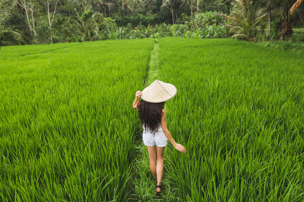 Visão traseira da mulher europeia morena andando em campos de arroz com chapéu de palha balinês tradicional em Ubud
 - Foto, Imagem