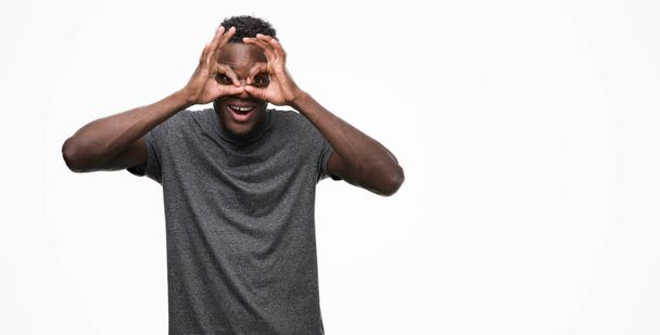 Jeune homme afro-américain portant un t-shirt gris faisant un geste correct comme des jumelles sortant la langue, les yeux regardant à travers les doigts. Expression folle
. - Photo, image