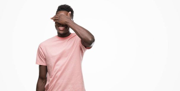 若いアフリカ系アメリカ人の男は微笑し、笑う顔驚きの目を覆っている手でピンクの t シャツを着てします。視覚障害者の概念. - 写真・画像