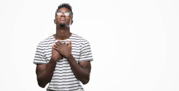 Young african american man wearing glasses and navy t-shirt smiling with hands on chest with closed eyes and grateful gesture on face. Health concept. - Photo, Image