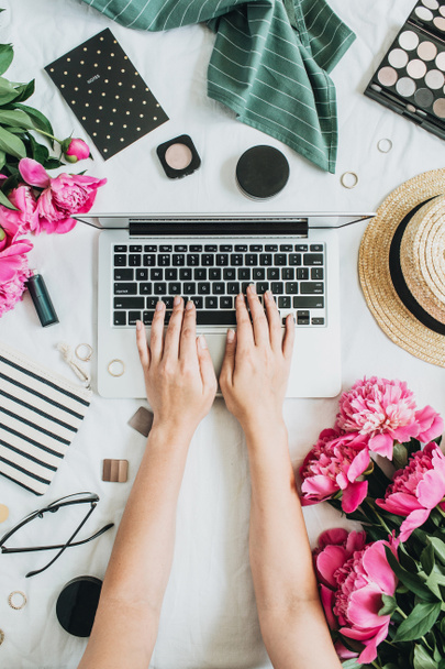 Woman working on laptop. Flat lay, top view female office desk workspace with pink peony flowers bouquet, accessories and cosmetics. Fashion, beauty or lifestyle blog concept. - Photo, Image