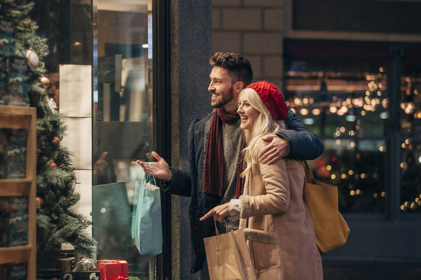 Side view of a couple doing some window shopping at christmas. The mid adult male has his arm around the mid adult female and they are talking about wats in the window. - Fotografie, Obrázek
