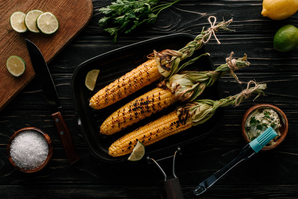 top view of griddle pan with grilled corn and lime slices surrounded by ingredients on wooden table  - Foto, afbeelding