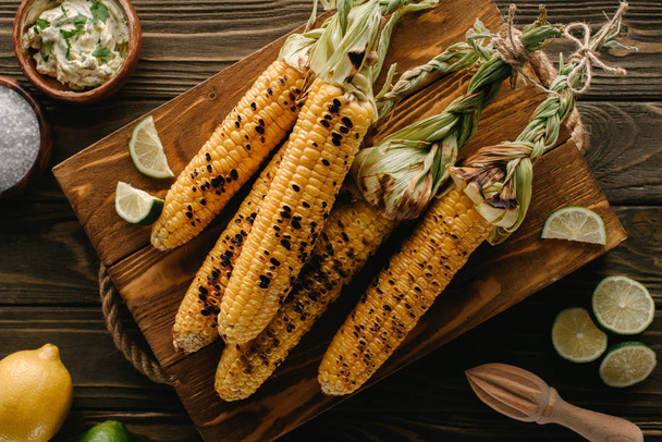 top view of cutting board with grilled corn, lime slices, salt, lemon, butter with parsley and squeezer on wooden table  - Zdjęcie, obraz