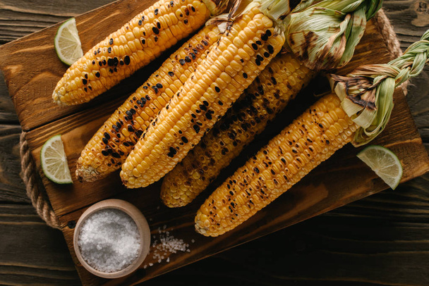 top view of cutting board with delicious grilled corn, lime slices and salt on wooden table  - Fotografie, Obrázek