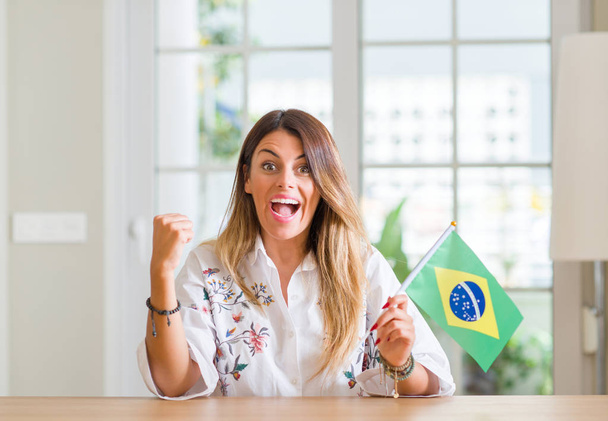 Νεαρή γυναίκα στο σπίτι εκμετάλλευση σημαία της Βραζιλίας ουρλιάζοντας περήφανος και τον εορτασμό νίκης και επιτυχίας πολύ ενθουσιασμένος, ζητωκραυγάζει συγκίνηση - Φωτογραφία, εικόνα