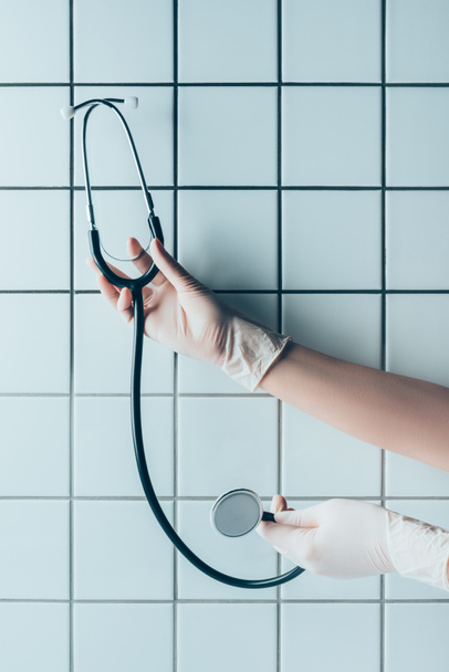 Schnappschuss von Arzt in Handschuhen mit Stethoskop vor gekachelter weißer Wand - Foto, Bild