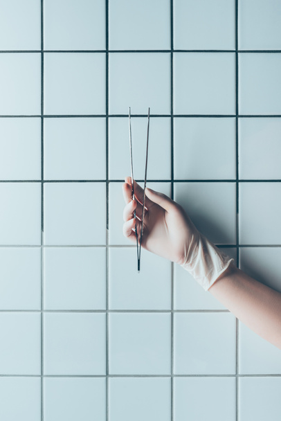 cropped shot of doctor in glove holding tweezers in front of tiled white wall - Foto, Imagem