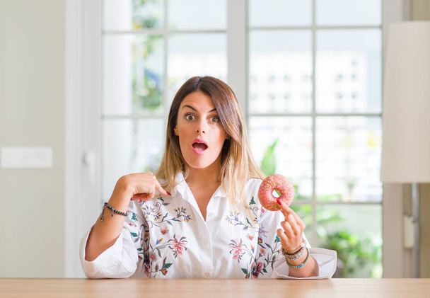 Young woman at home eating a doughnut with surprise face pointing finger to himself - Foto, Imagem