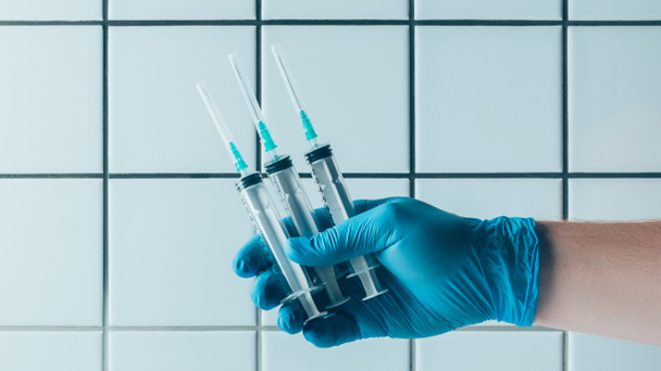 cropped shot of doctor in blue glove holding syringes in front of tiled white wall - 写真・画像
