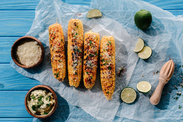 top view of grilled corn on baking paper with lime slices, squeezer, cream with parsley and spice  - Photo, Image