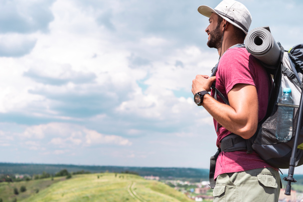 turista masculino en sombrero con mochila mirando al prado de verano
 - Foto, Imagen