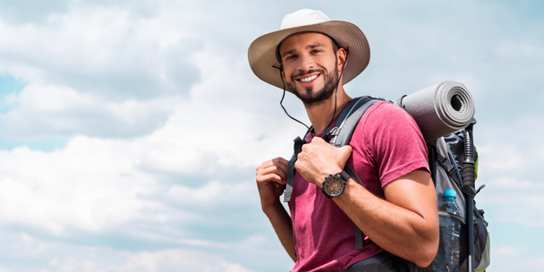 viajante sorridente em chapéu com mochila e tapete turístico, com fundo céu nublado
 - Foto, Imagem