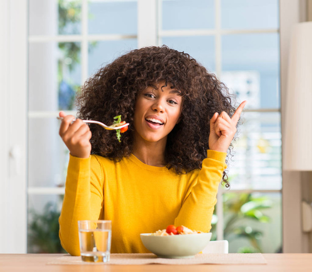 African american vrouw eten pastasalade thuis verrast met een idee of vraag wijzende vinger met blij gezicht, nummer één - Foto, afbeelding