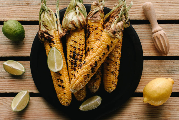 top view of grilled corn on plate surrounded by lime slices and lemon with squeezer on wooden table  - Fotografie, Obrázek
