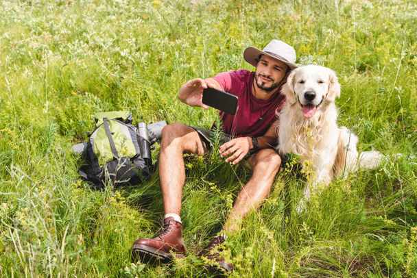 traveler in hat taking selfie on smartphone with dog on summer meadow - Foto, Bild