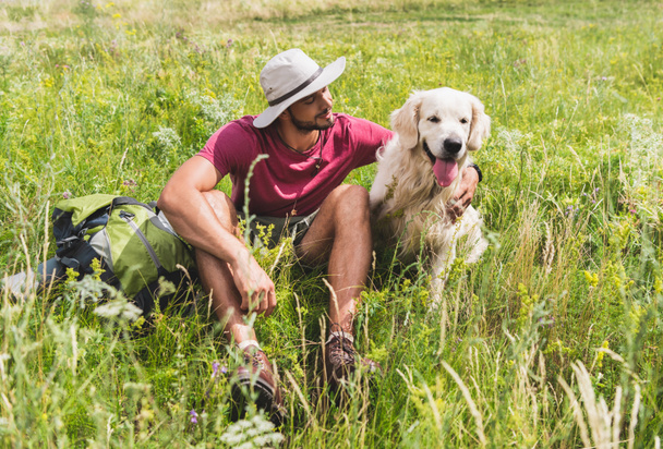 traveler in hat sitting with golden retriever dog on green meadow - Photo, image