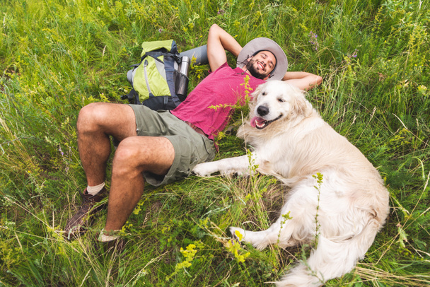 top view of smiling traveler and golden retriever dog lying on green grass with backpack - Foto, imagen