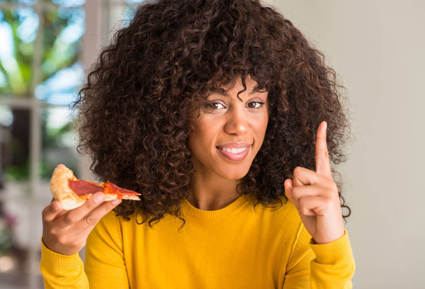 African american woman ready to eat pepperoni pizza slice surprised with an idea or question pointing finger with happy face, number one - Photo, image