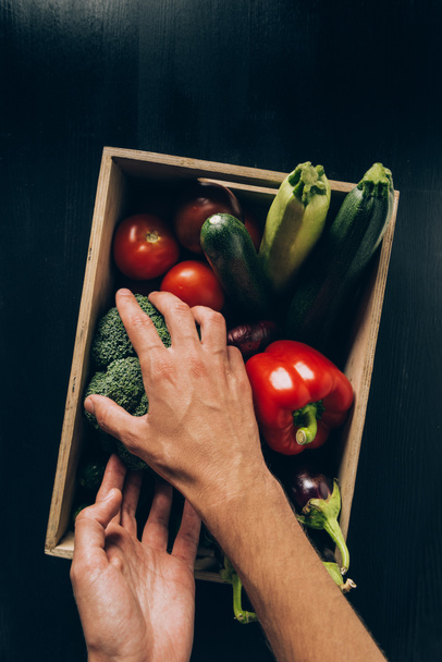 cropped image of man taking broccoli from wooden box with different vegetables - Foto, afbeelding