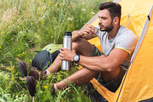 турист сидит в желтой палатке и пьет кофе из термоса
 - Фото, изображение