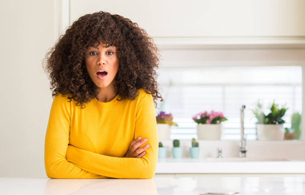 Donna afroamericana che indossa maglione giallo in cucina spaventata e scioccata con espressione a sorpresa, paura e viso eccitato
. - Foto, immagini