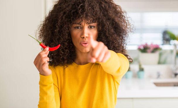 Africká americká žena jíst hot chili paprika ukazující prstem na kameru a na vás, rukou znamení, pozitivní a sebevědomý gesto z přední - Fotografie, Obrázek