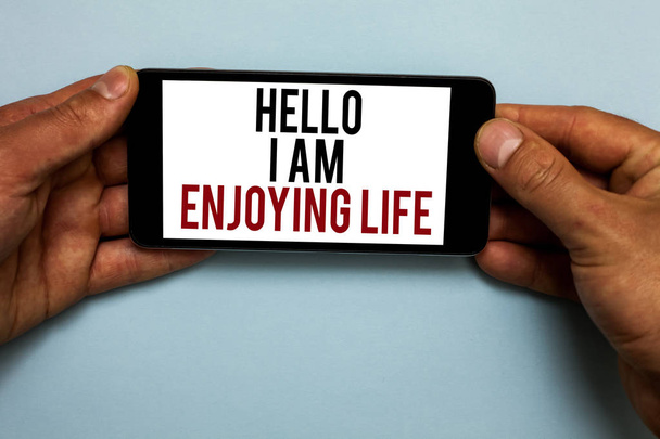 Word writing text Hello I Am Enjoying Life. Business concept for Happy relaxed lifestyle Enjoy simple things Human hand hold smartphone with red and black letters on shadow blue floor - Photo, Image