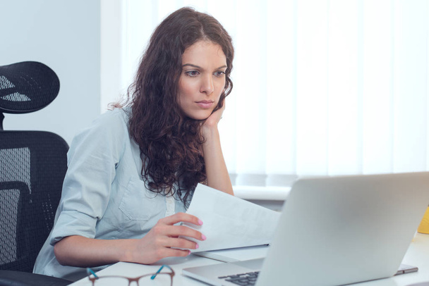 Успішна серйозна бізнес-леді в офіційному одязі сидить за білим столом в сучасному офісі і читає на ноутбуці, копіює простір
 - Фото, зображення