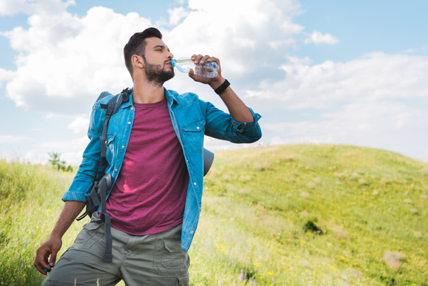 handsome traveler with backpack drinking water on summer meadow - Photo, Image