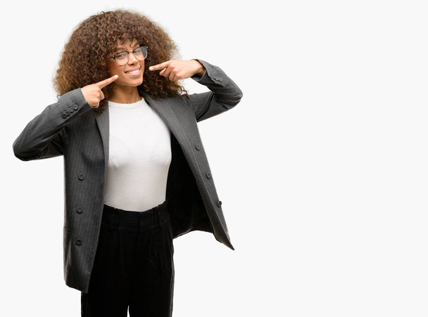 Donna d'affari afroamericana con gli occhiali sorridente fiducioso mostrando e indicando con i denti delle dita e la bocca. Concetto sanitario
. - Foto, immagini