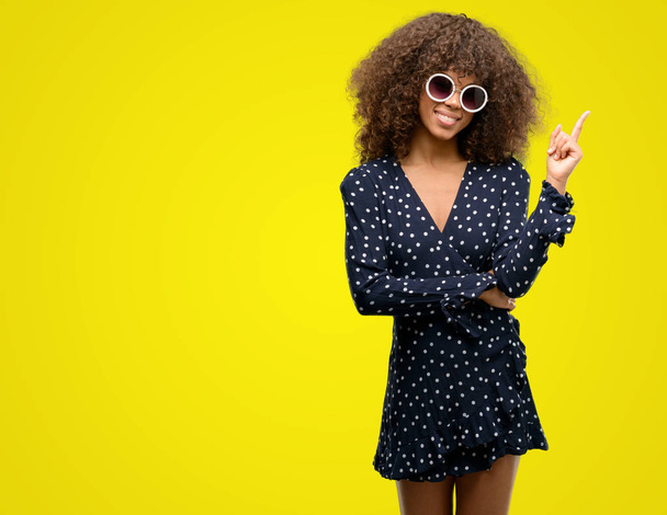 Африканская американка в солнечных очках и летнем платье очень счастлива, указывая рукой и пальцем в сторону
 - Фото, изображение
