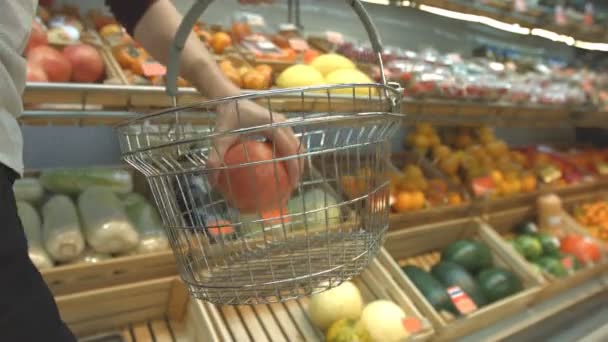 Cesta de frutas en el supermercado.Las manos de los hombres llevan una cesta de comestibles en el supermercado y ponen diferentes frutas en ella: granada, melón, naranjas, mandarinas. En los estantes hay muchas otras frutas y verduras: caquis, fresas, sandías
,  - Metraje, vídeo