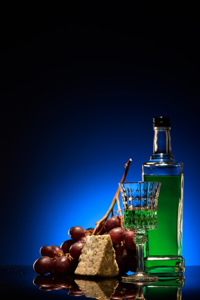 Close-up shot van absint met druiven en dorblu kaas op spiegelend oppervlak op donker blauwe achtergrond - Foto, afbeelding