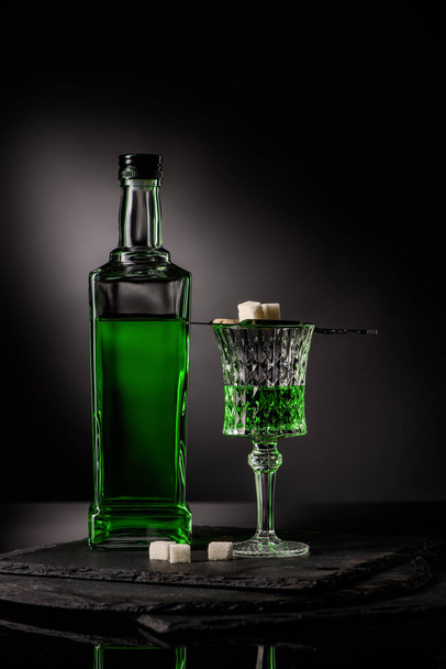 крупным планом стекло и бутылка абсента с ложкой и кубиками сахара на темном фоне
 - Фото, изображение