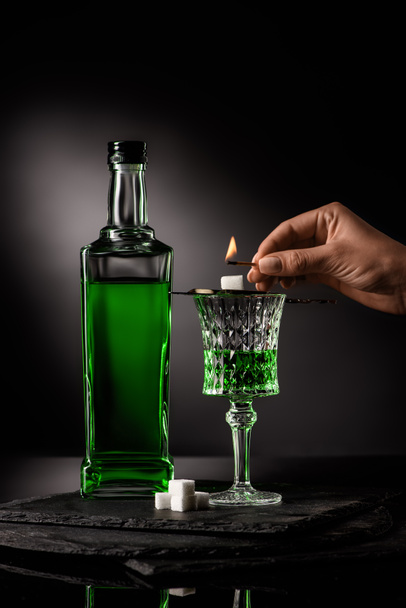 cropped shot of woman holding match over spoon with sugar cube on absinthe glass on dark background - 写真・画像