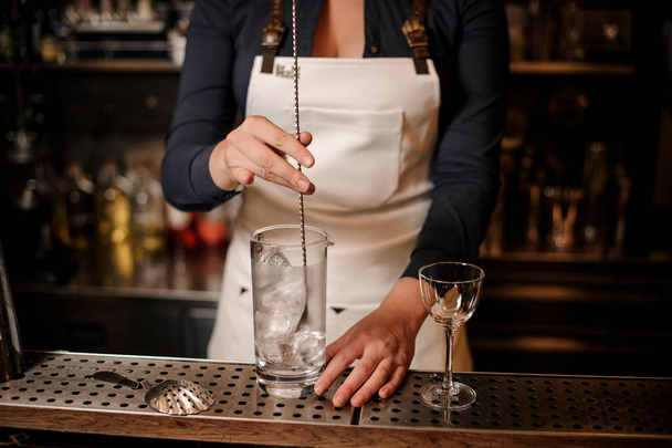 Female bartender in the white apron mixing an ice cubes in the glass at the bar counter - Foto, Imagen