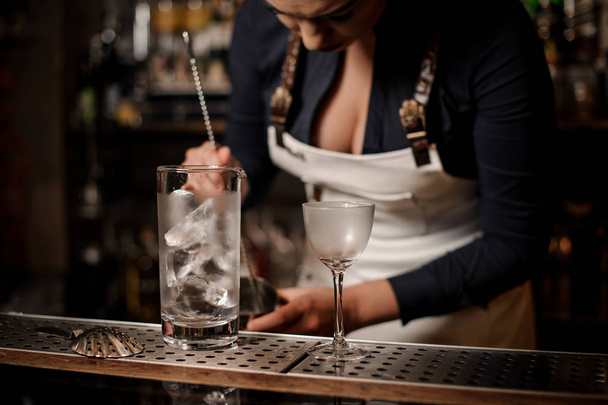 Glass with ice on the background of female bartender stirring cocktail on the bar counter - Foto, Imagen