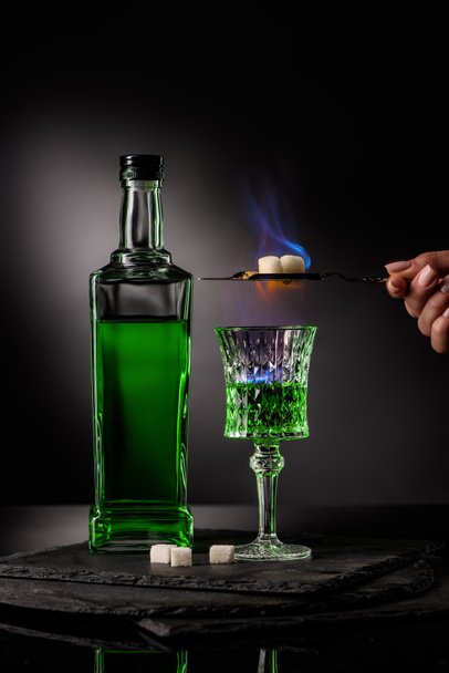 cropped shot of woman holding spoon with sugar cubes over burning absinthe in glass on dark background - 写真・画像