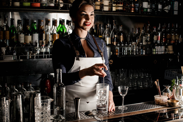 Female bartender in the white apron stirring an alcoholic drink in the glass with ice cubes at the bar counter - Foto, afbeelding