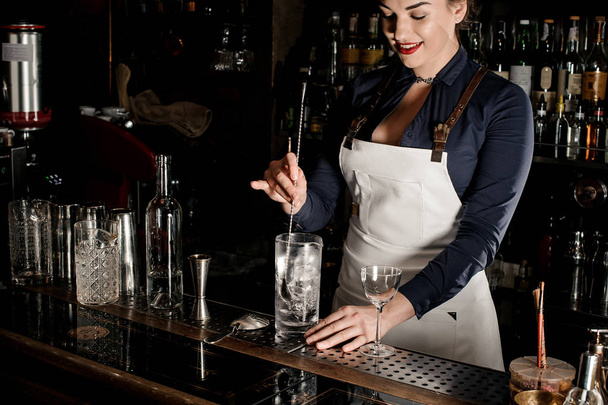 Smiling female bartender in the white apron stirring an alcoholic drink in the glass with ice cubes at the bar counter - Zdjęcie, obraz
