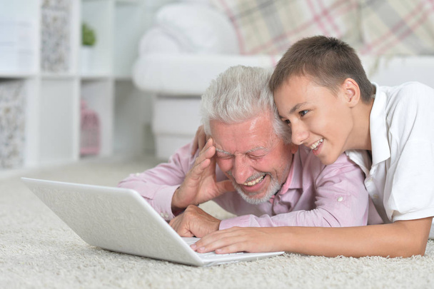 家の床に横たわっている間ノート パソコンを使用する孫と祖父 - 写真・画像