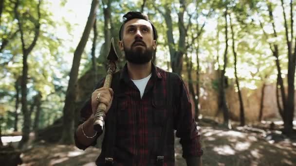 Walking bearded lumberjack with axe. Man in a cap walks through the woods in search of the tree - Metraje, vídeo