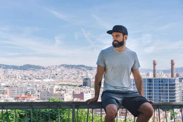 Bearded muscular hipster man model wearing gray blank t-shirt and a black baseball cap with space for your logo or design in casual sport urban style.Panoramic view of modern city on the background. - Fotó, kép