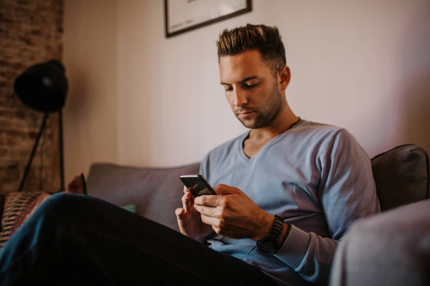 Handsome man using mobile smartphone for surfing web internet and sending audio message. Man using gadget at modern apartment. - Fotografie, Obrázek